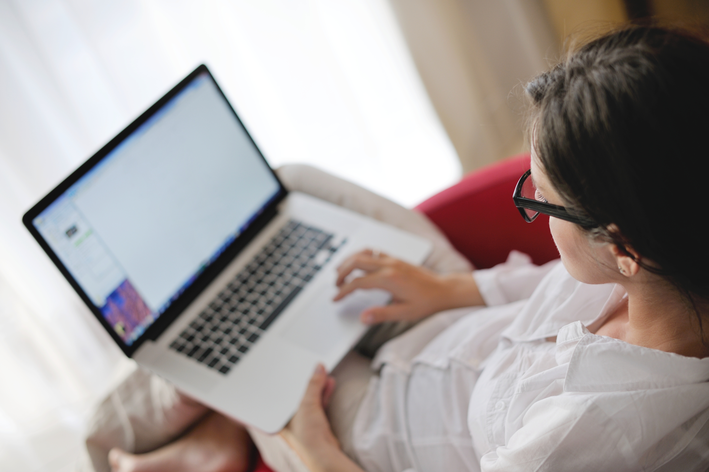 A woman wearing glasses and a white blouse sits relaxed on a red sofa, using a laptop with its screen visible. she appears focused on her work in a softly lit room.