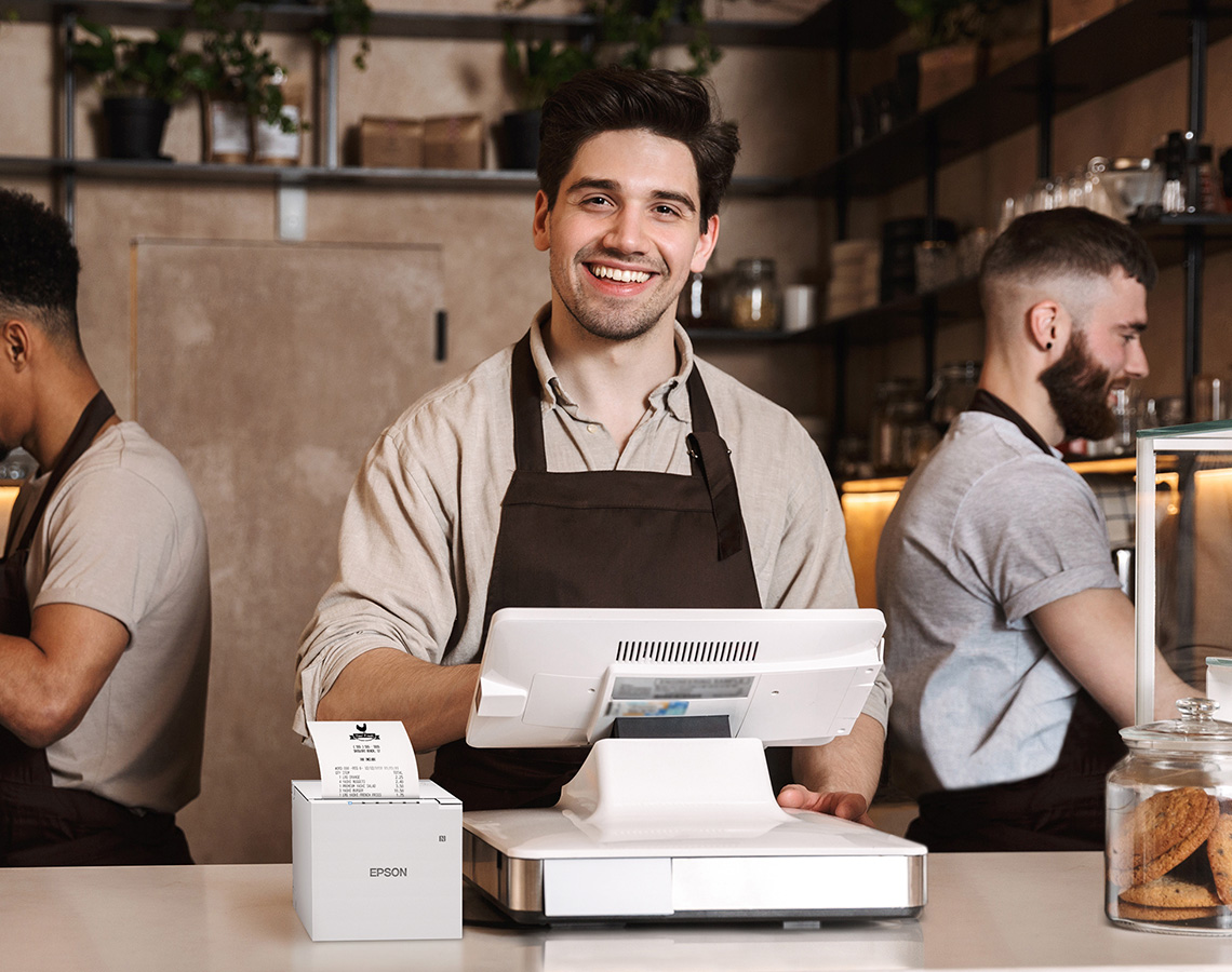 A man standing at a register next to an Epson M-Series printer.