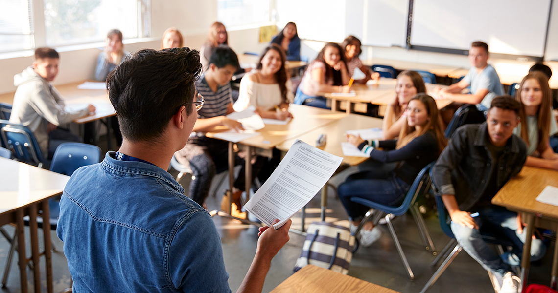 Teacher holding printed lecture notes in front of high school classroom.