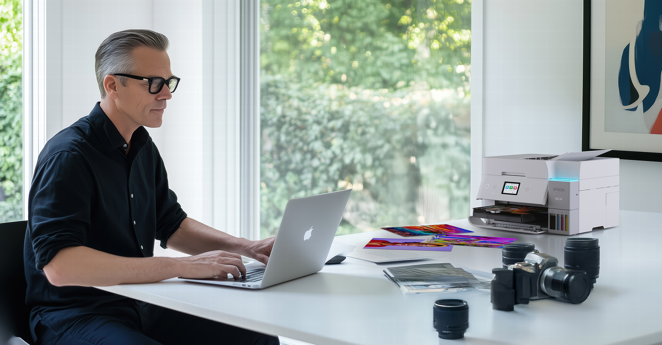 Man working on a laptop in a bright, modern home office with an EcoTank printer on the table, surrounded by camera equipment and vibrant photo prints.