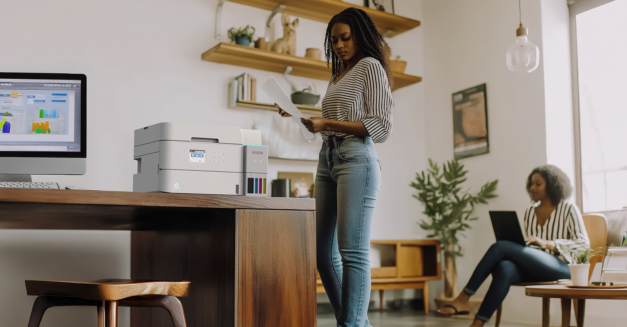 Two women working in a modern office; one stands at a desk reviewing printed documents next to an EcoTank printer, while the other sits in the background using a laptop.