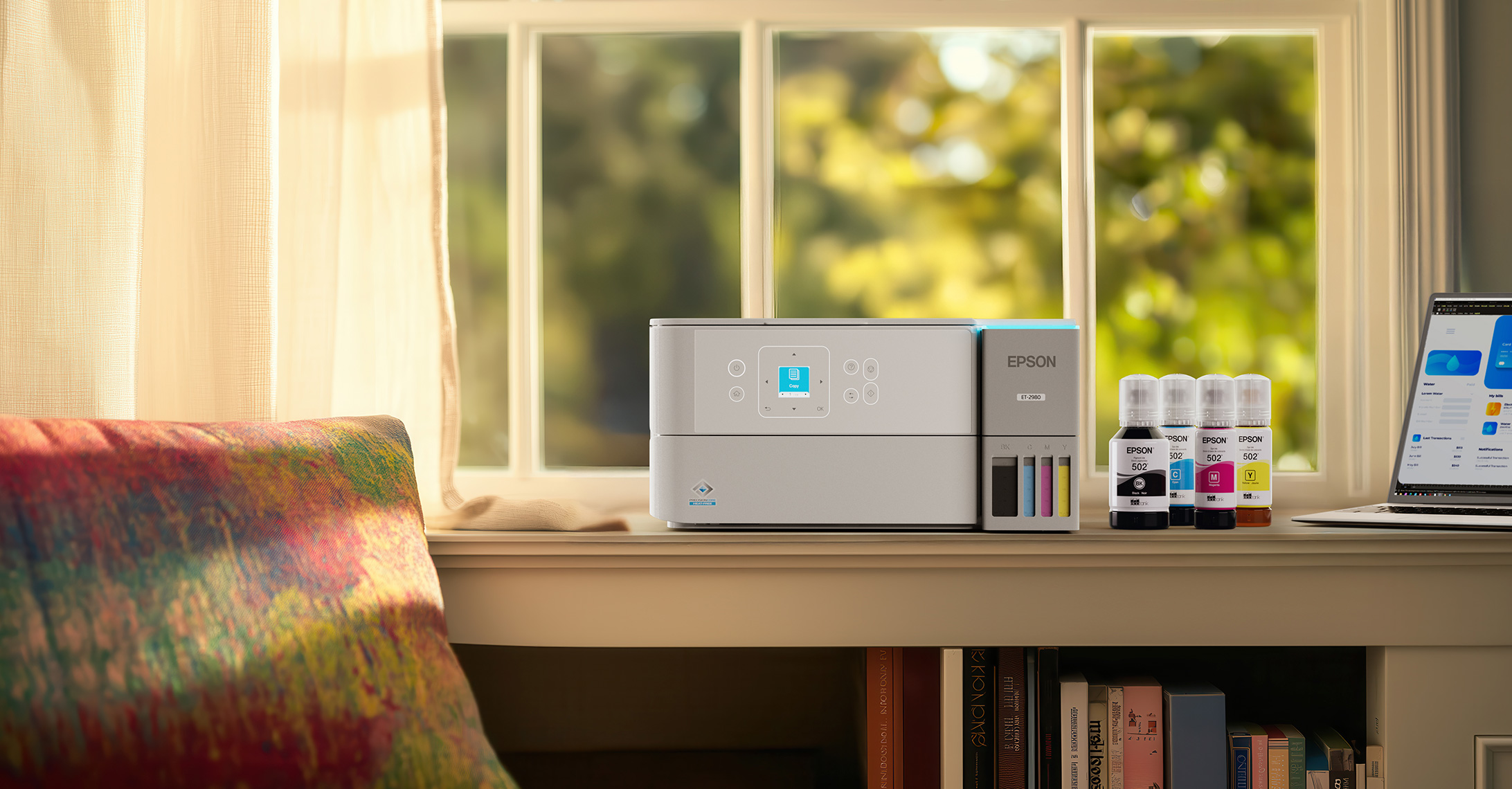 EcoTank printer on a bookshelf beside ink bottles and a laptopwith sunlight streaming through a nearby window.