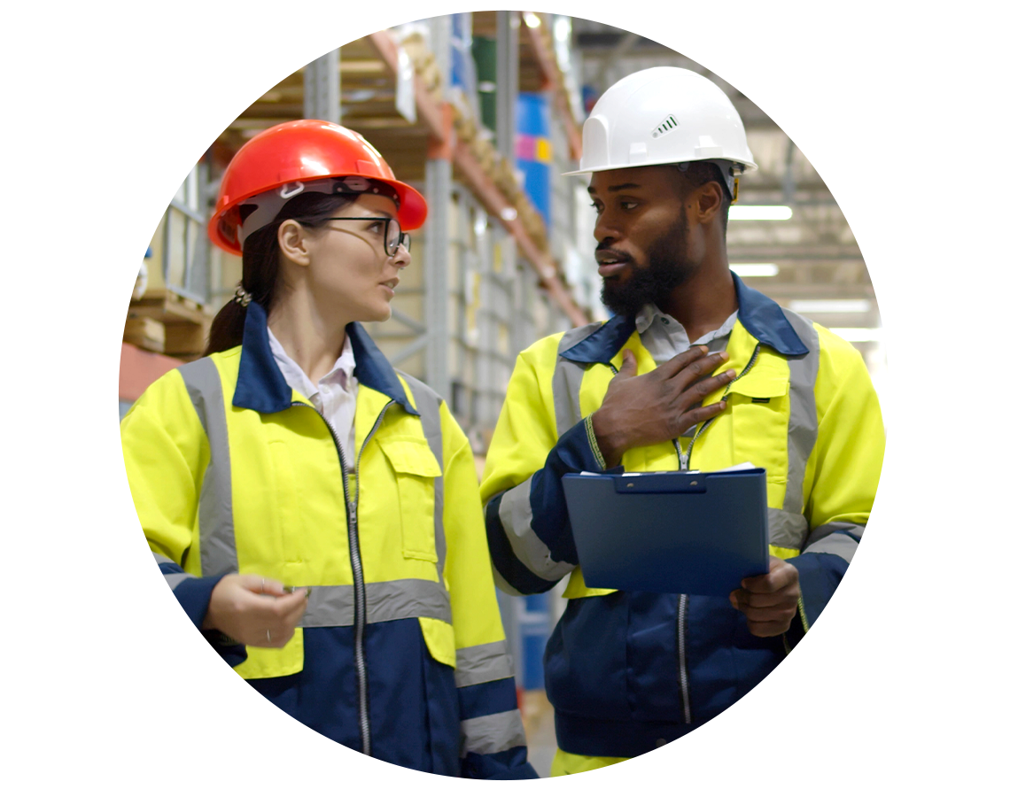 Two chemical warehouse employees talking and walking through rows of palettes.