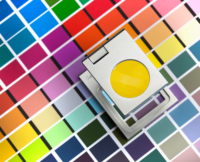 A densitometer tool rests on a colorful swatch of printed color bars, used for calibrating and measuring print color accuracy in a graphic design setting.