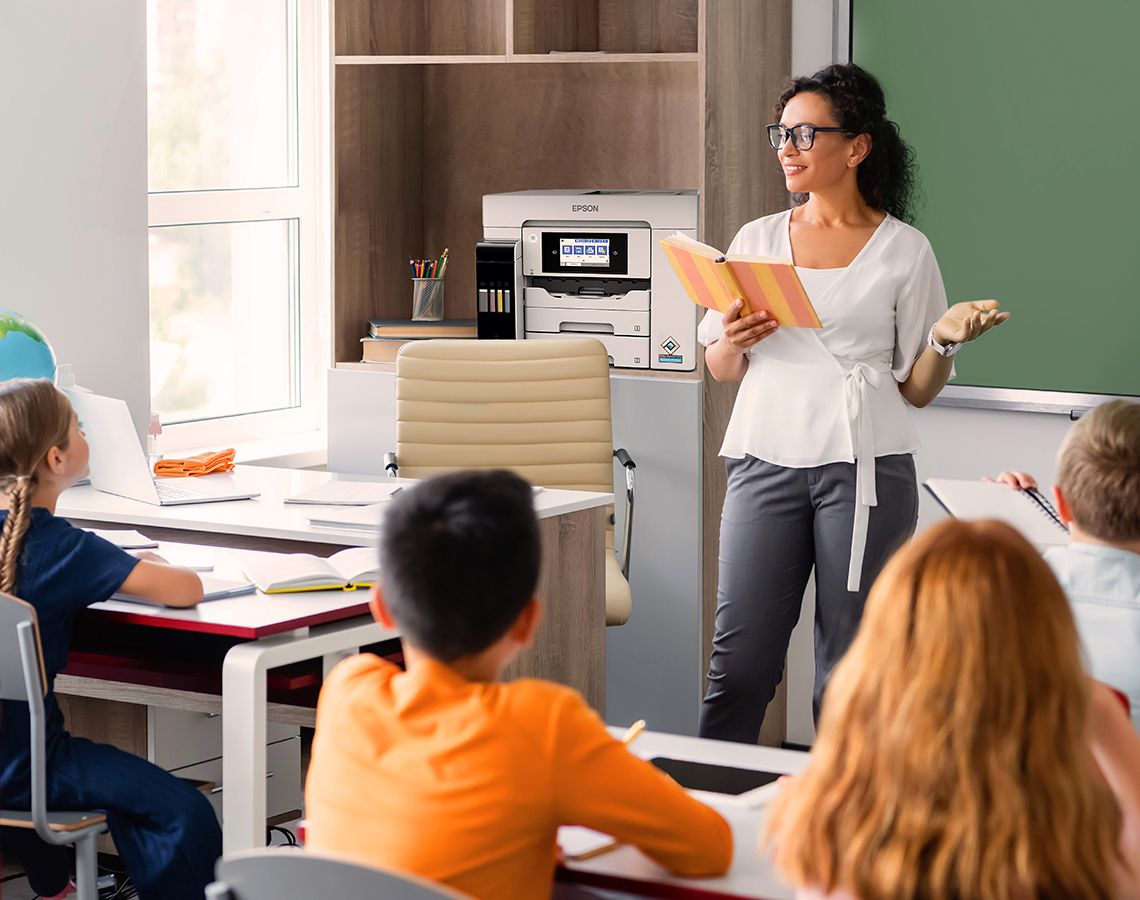 Teacher reading from book to elementary school students with Epson ST-C5500 printer on shelf.