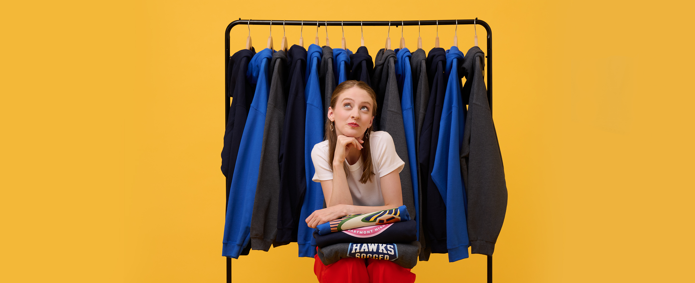 Woman leaning on stack of printed hoodies in front of clothing rack.